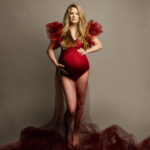 Beautiful woman wearing a red body suit with ruffled sleeves and a red net veil covering her pregnant bump and it flows over her legs she has a powerful look by norfolk photographer